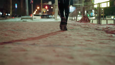 first-person view of a woman wearing black leather trousers and black boots, walking at night on a snowy street. blurry city lights and cars are visible in the background