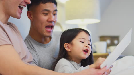 Familie-Mit-Zwei-Vätern-Im-Bett-Zu-Hause,-Die-Ihrer-Tochter-Eine-Geschichte-Vorlesen