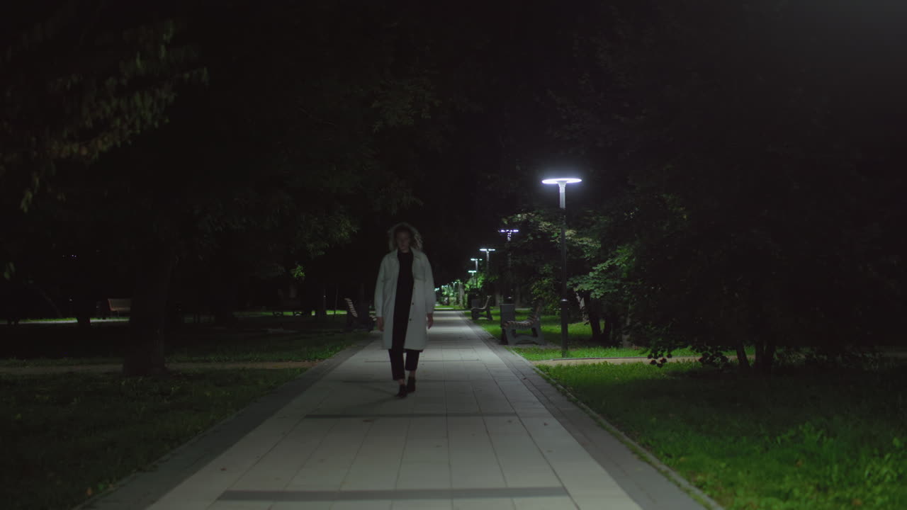 Woman in white coat walks alone along nighttime walkway under soft lamplight, foliage framing serene park ambiance, sense of solitude and reflection, gentle steps echoing in quiet environment