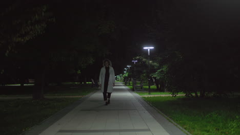 woman in white coat walks alone along nighttime walkway under soft lamplight, foliage framing serene park ambiance, sense of solitude and reflection, gentle steps echoing in quiet environment