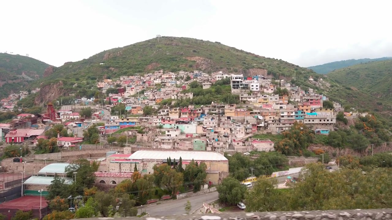 Kota di lereng bukit dengan bangunan tempat tinggal padat, rumah-rumah berwarna-warni, pegunungan di sekitarnya, dan vegetasi hijau di Cuernavaca, Meksiko di bawah cahaya siang yang mendung