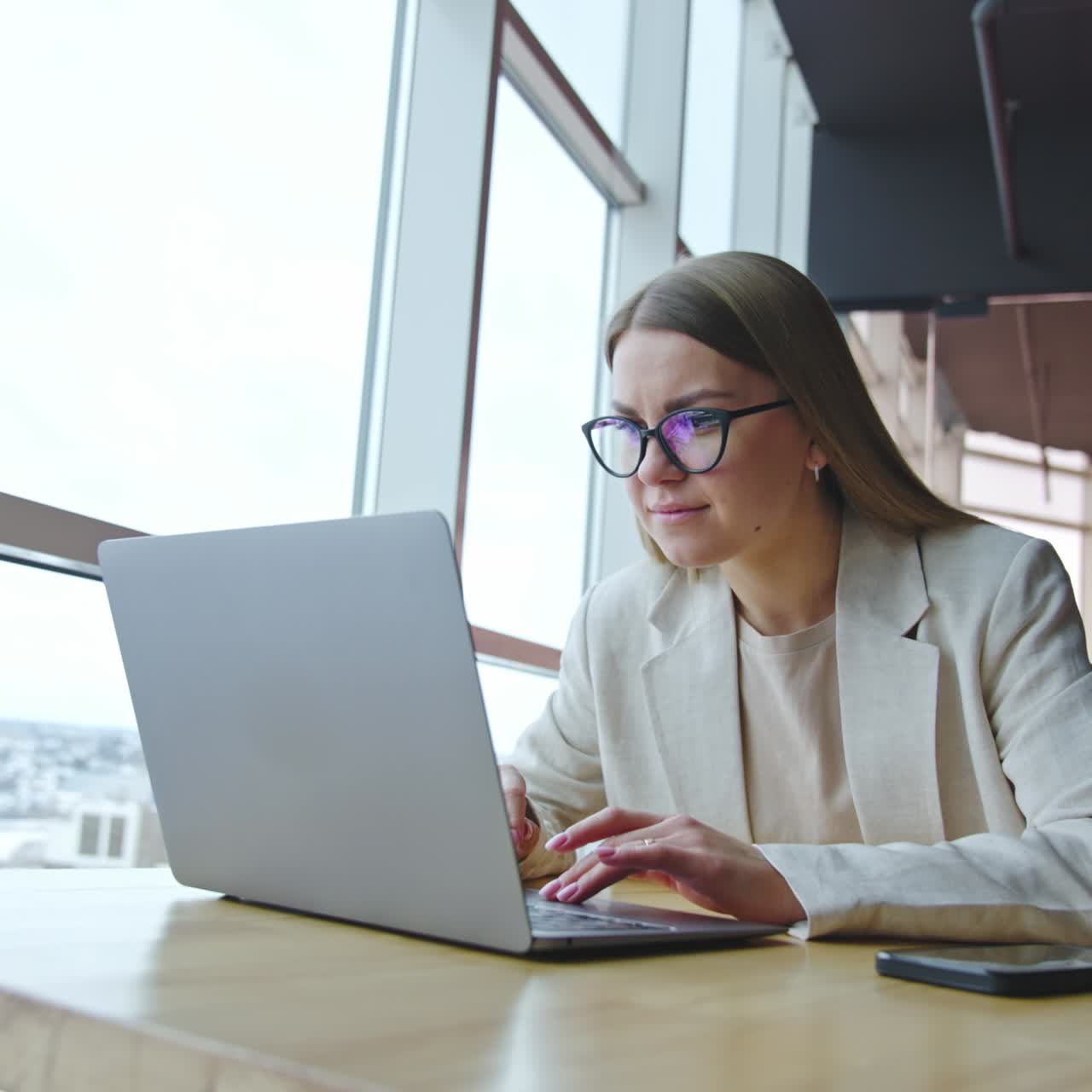 Young woman works on her laptop sitting at the desk. Lady in glasses looking intently at the screen. Workplace at the panoramic window