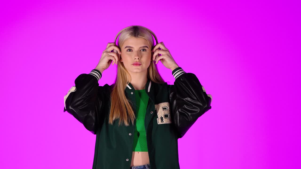 una joven rubia escuchando música con auriculares y bailando, retrato de estudio con fondo rosa