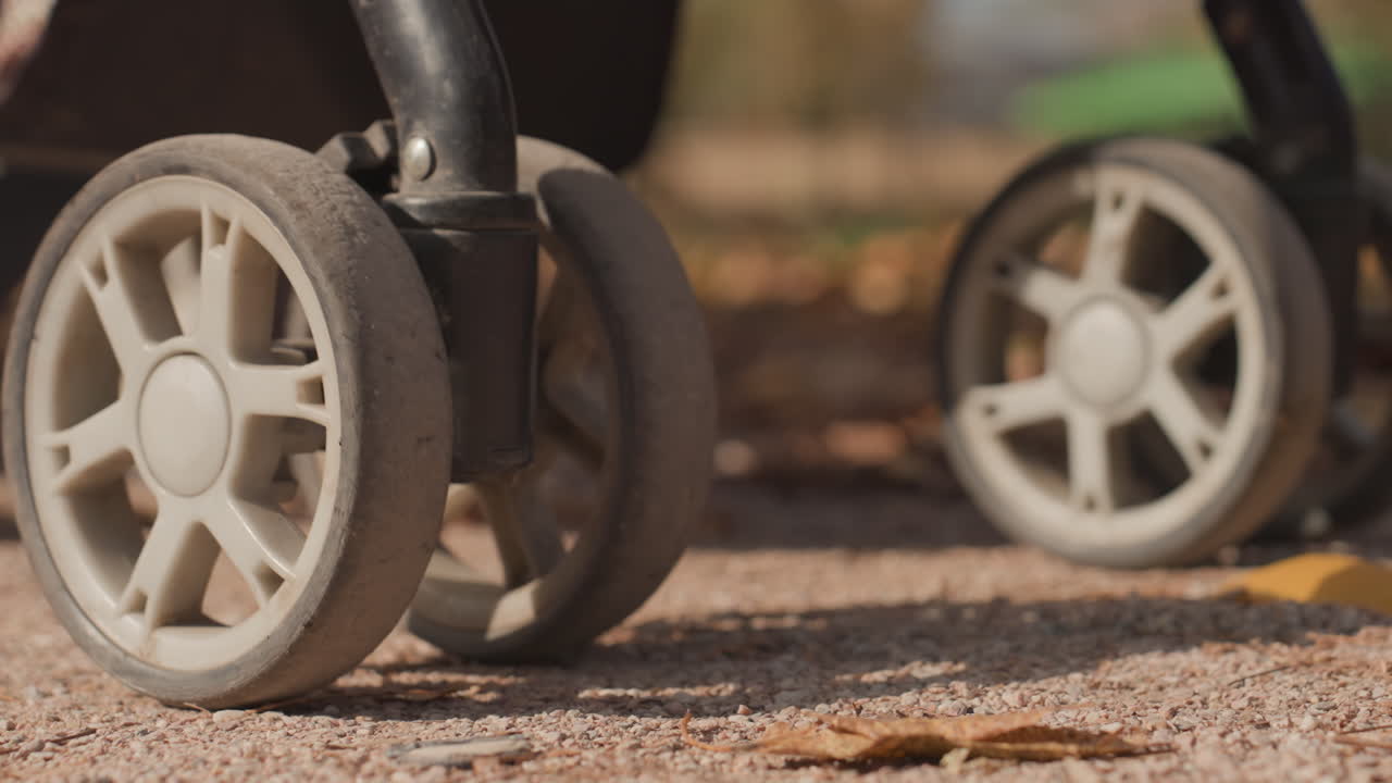 Primo piano delle ruote del passeggino sulla ghiaia, battistrada in gomma dettagliato e rotazione della ruota. Macchie di fango e telaio strutturato, movimento costante lungo il sentiero del parco, luce solare e ombra tenui, simbolismo del matrimonio condiviso.