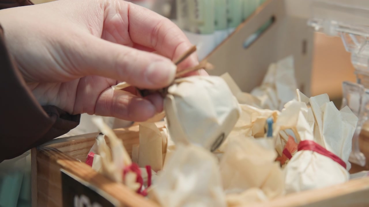 Hand selecting wrapped soap from wooden crate at artisan market, twine tied packaging and ribbon accents, closeup of fingers picking creamy bar, vendor stall backdrop and handcrafted boutique vibe