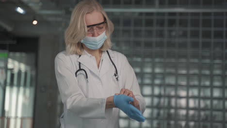 a blonde, middle aged female doctor in a facemask puts on surgical gloves