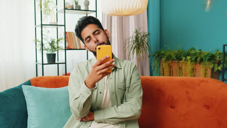Indisch-arabischer-Junger-Mann-Sitzt-Auf-Dem-Sofa-Und-Nutzt-Sein-Smartphone,-Um-Nachrichten-In-Einer-Social-Media-Anwendung-Zu-Teilen
