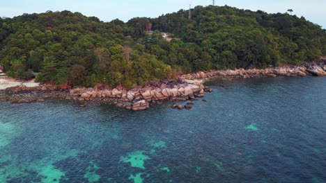 Capturing-the-beauty-of-a-tropical-island,-turquoise-waters-and-rocky-coastline,-this-aerial-view-shows-vibrant-blue-and-green-hues-against-rugged-cliffs-and-shores