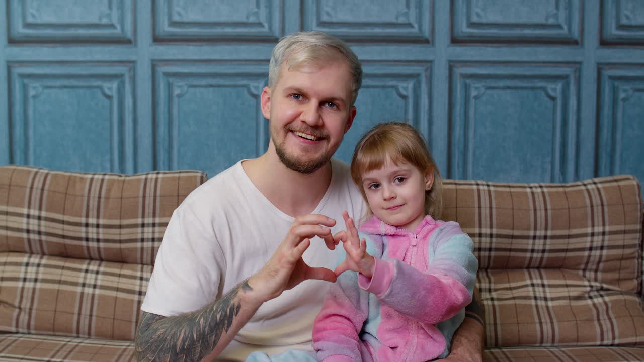 Far og datter sidder i tøj på sofaen, smiler og laver et hjerte-tegn nær brystet.