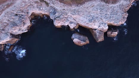 Top-down-aerial-view-of-waves-splash-against-rocky-seashore,-background