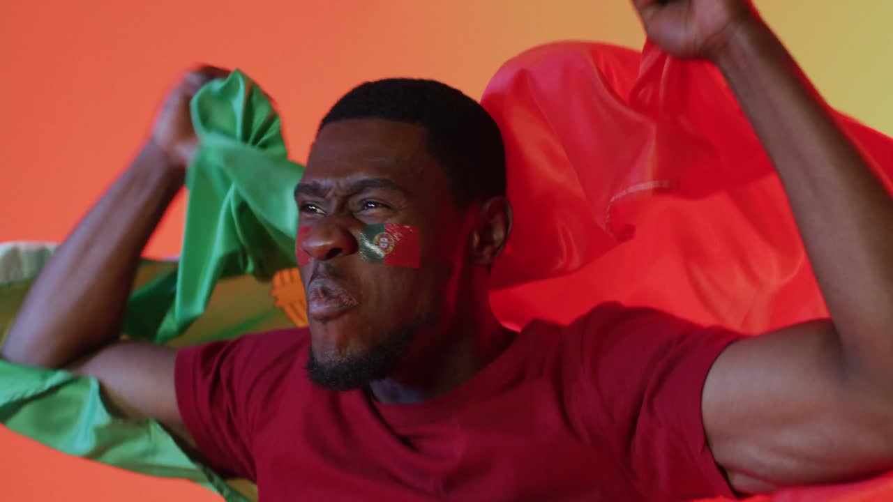 Happy african american male football supporter with flag of portugal over pink lighting