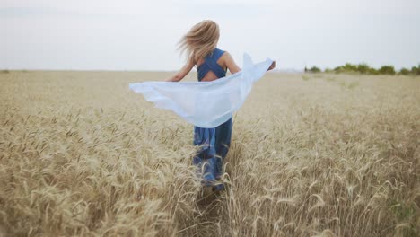 Rückansicht-Einer-Attraktiven-Jungen-Frau-In-Einem-Langen-Blauen-Kleid,-Die-Durch-Ein-Goldenes-Weizenfeld-Läuft-Und-Einen-Schal-In-Ihren-Händen-Hält.-Die