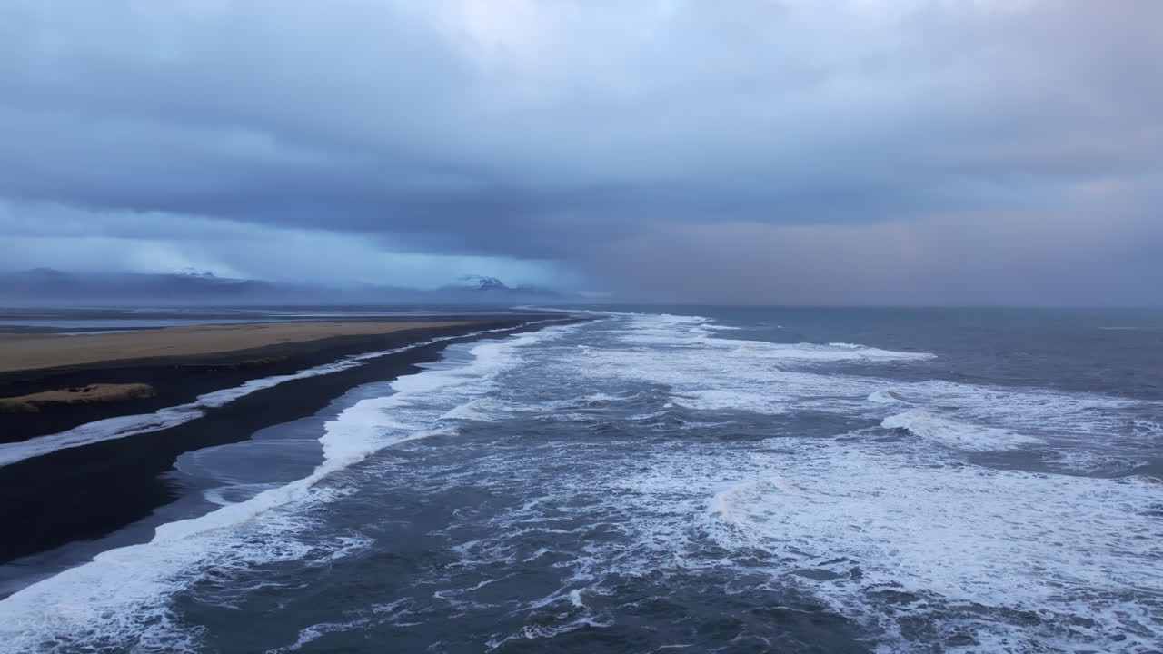 Kraftige bølger slår inn mot en svart sandstrand under mørke, illevarslende skyer, og skaper en dramatisk kystscene som viser naturens rå kraft, Island, flyfoto