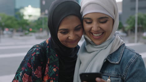 portrait of young muslim women posing daughter kisses mother on cheek taking selfie photo using smartphone camera technology in urban city background