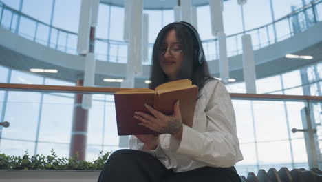 young girl wearing headphones smiles as she dances slightly while reading book seated in bright indoor mall space, surrounded by glass railings, plants, and modern architectural elements