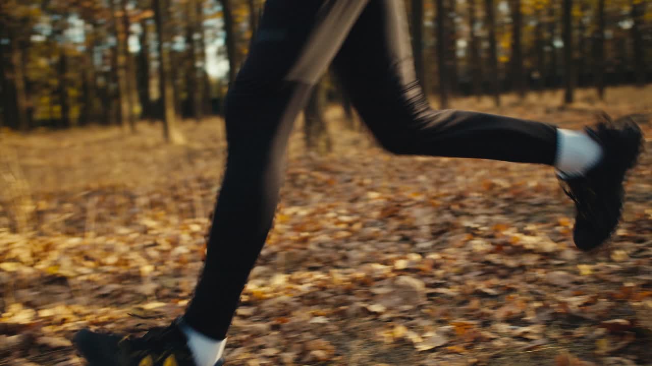 Confident sporty man with curly hair in a black sports uniform runs through the forest with fallen yellow leaves on a sunny autumn day. Male athletes in black sportswear run between trees with fallen brown leaves in the autumn morning