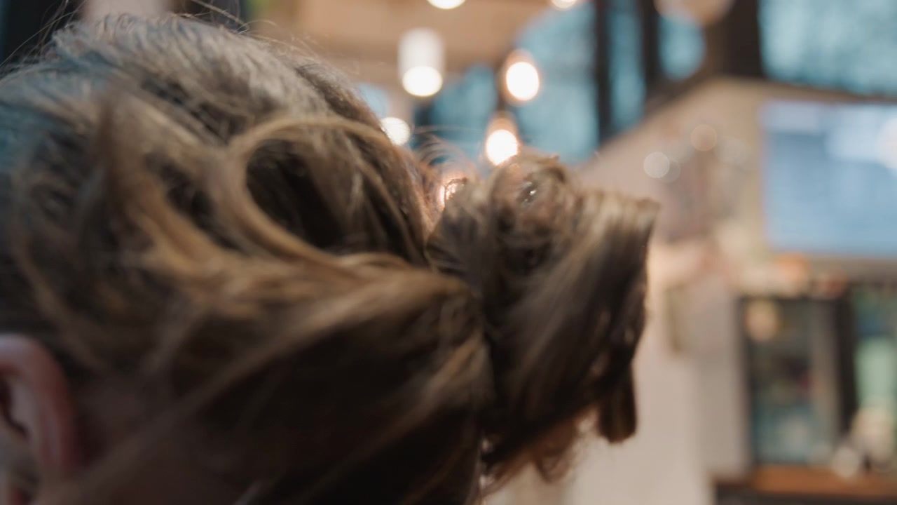 Young caucasian woman tying bun in cozy cafe, closeup hands twisting hair near warm bokeh lights, casual sweater with thumbholes, behindthescenes calm, natural texture, intimate grooming routine