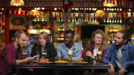 a large group of friends in the bar sitting at the table laughing and chatting drinking beer.