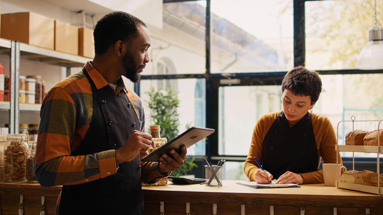 Tapping tablet, man wearing apron checking orders at wooden counter, coworker pausing to take notes