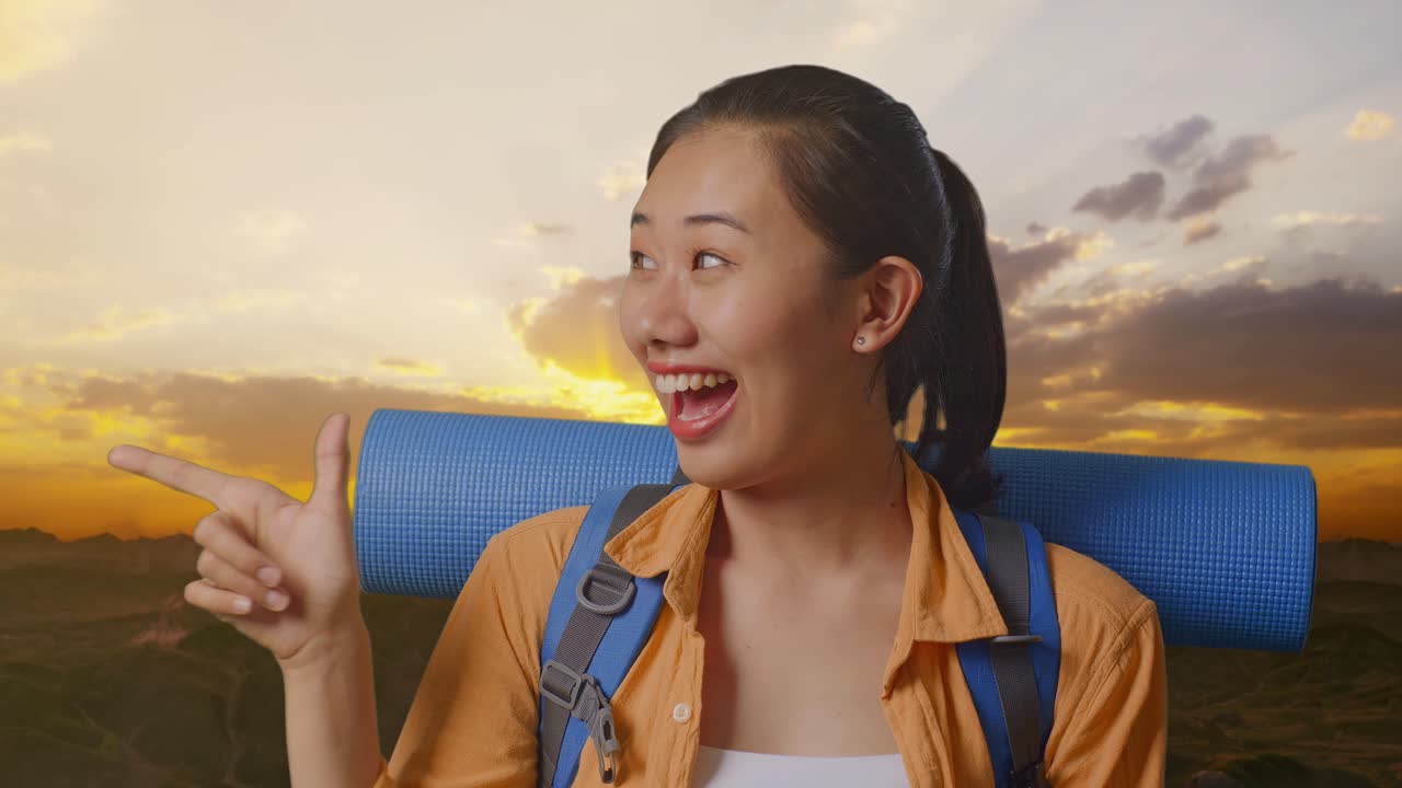 близкий взгляд на азиатскую туристку с альпинистским рюкзаком, улыбающаяся и указывающая в сторону, стоя на вершине горы во время захода солнца