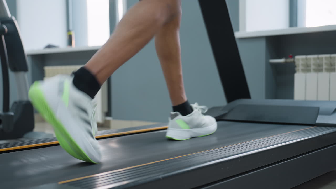 leg view of man in black shorts and white sneakers walking briskly on treadmill in spacious gym, sunlight streaming through windows onto tiled floor, adjacent cardio machines