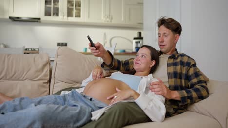 Happy-man-in-a-plaid-shirt-with-his-pregnant-wife-lying-on-the-sofa-and-watching-TV-during-their-relaxation-at-home