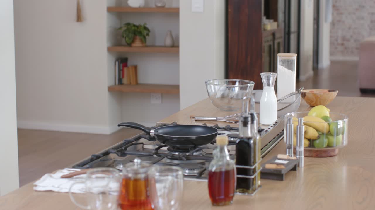 La telecamera si avvicina delicatamente all'isola della cucina, rivelando una padella sul piano cottura a gas, una teiera e della frutta.