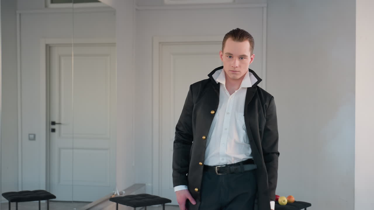 man showcasing fashion outfit walking forward while flaunting long black coat in modern studio with apples on stool and candle stand reflected in wall mirror, highlighting style and movement