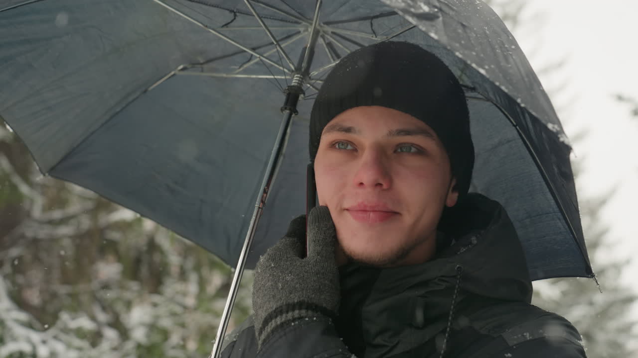 Homme souriant sous un parapluie, parlant au téléphone, conversation joyeuse dans une forêt enneigée, portant un bonnet tricoté et une veste matelassée, main gantée tenant le téléphone, parapluie mouillé parsemé de flocons, naturel