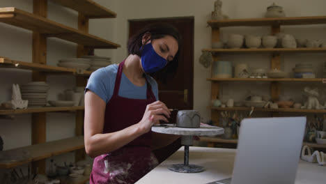 female caucasian potter wearing face mask using ribbon tool to create designs on pot while having a