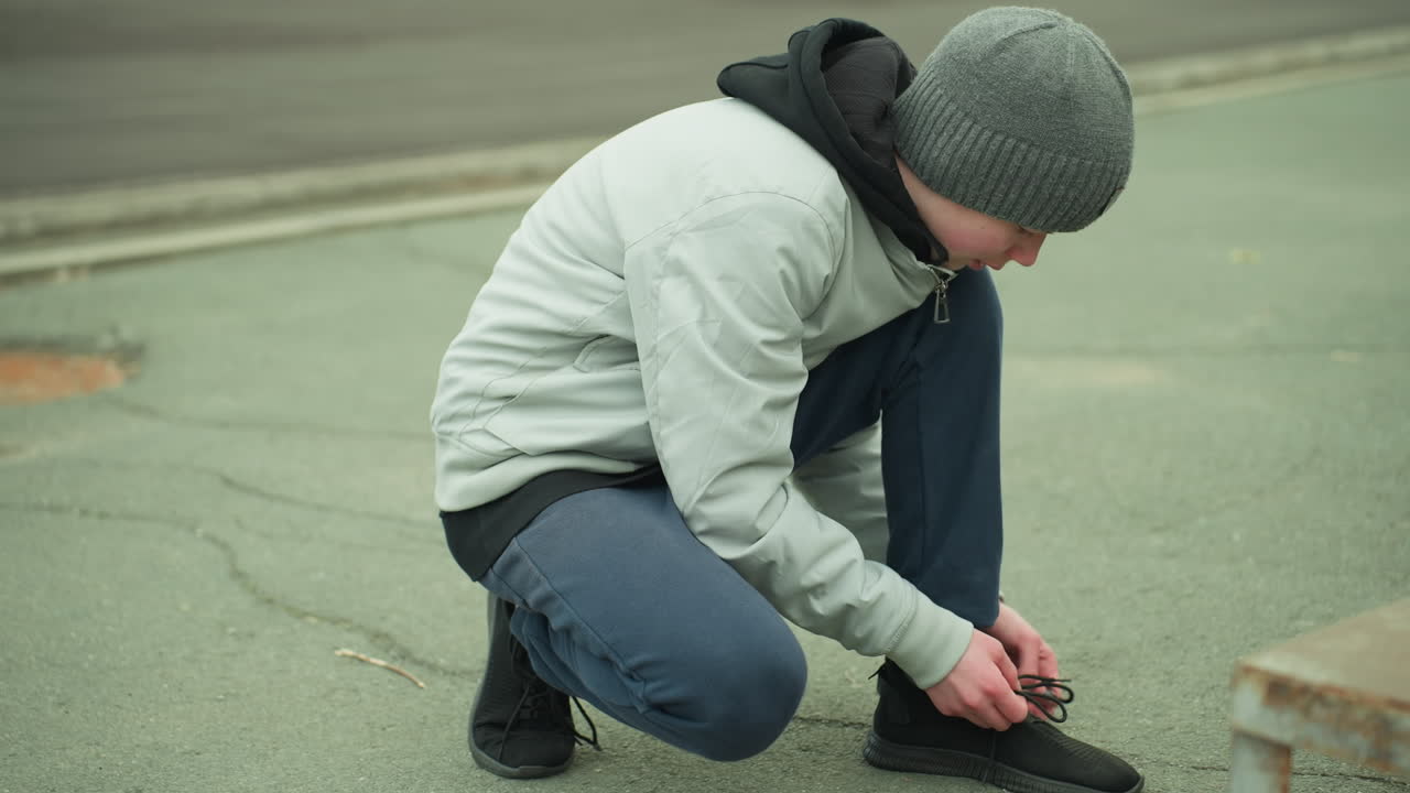A young boy kneeling down tying his shoelace on a paved road, wearing a gray beanie and jacket, the background is slightly blurred with a minor pothole