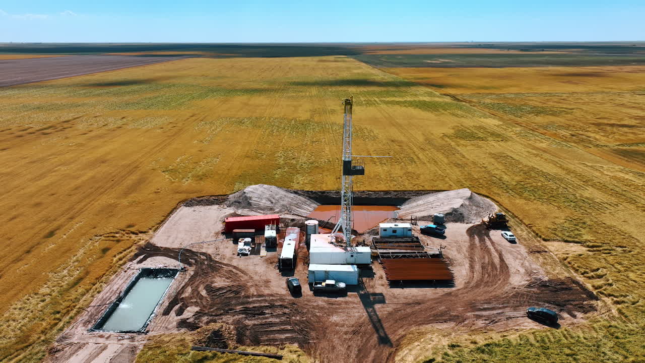 Oil production site in the vast field. Derrick in the middle of the location. Drone footage above the plantation on sunny daytime.
