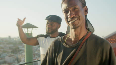 Front-view-of-gay-man-standing-on-rooftop-and-squinting-in-sun-with-his-boyfriend-dancing-behind