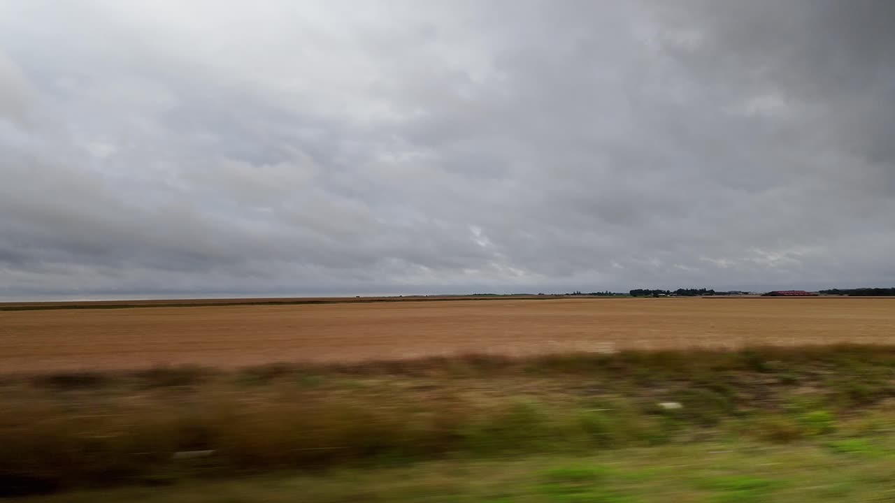 Point of view shot from a moving train or car, looking out at vast, flat agricultural fields in the French countryside under an overcast, cloudy sky