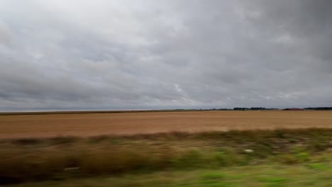 point of view shot from a moving train or car, looking out at vast, flat agricultural fields in the french countryside under an overcast, cloudy sky