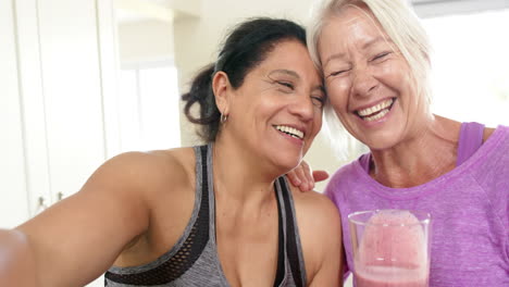 Dos-Mujeres-Mayores-Felices-Y-Diversas-Con-Un-Cóctel-Haciendo-Selfie-Riéndose-En-La-Cocina,-Cámara-Lenta