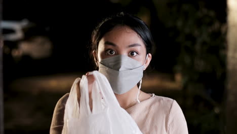 surprised asian woman wearing mask receiving a package in plastic bag, front shot