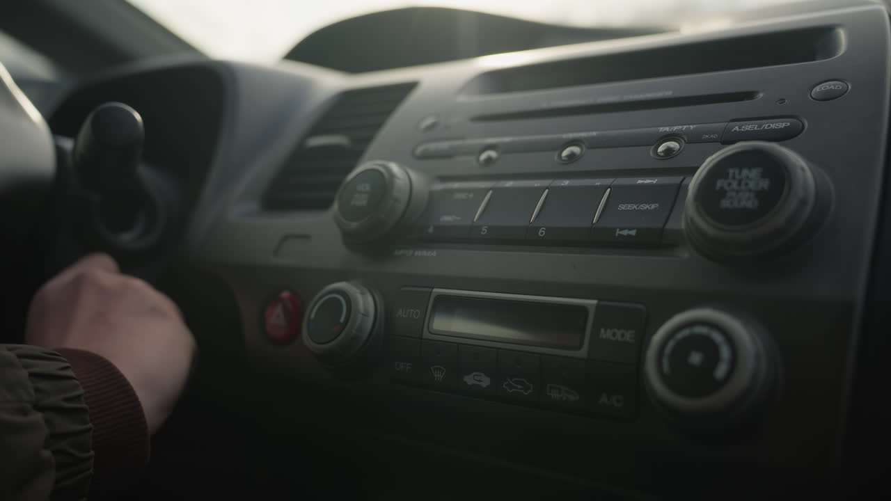 close up of driver hand turning off ignition inside parked vehicle, dashboard partially illuminated with visible stereo system and ventilation controls in foreground under natural daylight
