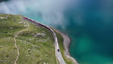 Train-moving-on-railroad-tracks-at-Bernina-Glacier-side-of-a-lake-in-Switzerland,-scenic-aerial-tracking-shot-at-Bianco-lake