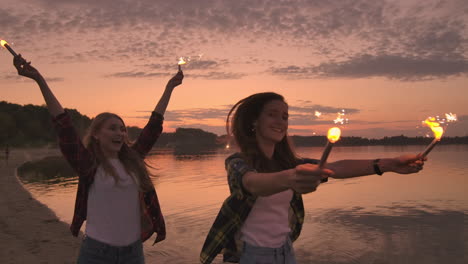 felices chicas sonrientes corriendo por la playa con chispas por la noche. sostengan las luces brillantes y rían estudiantes jóvenes despreocupados