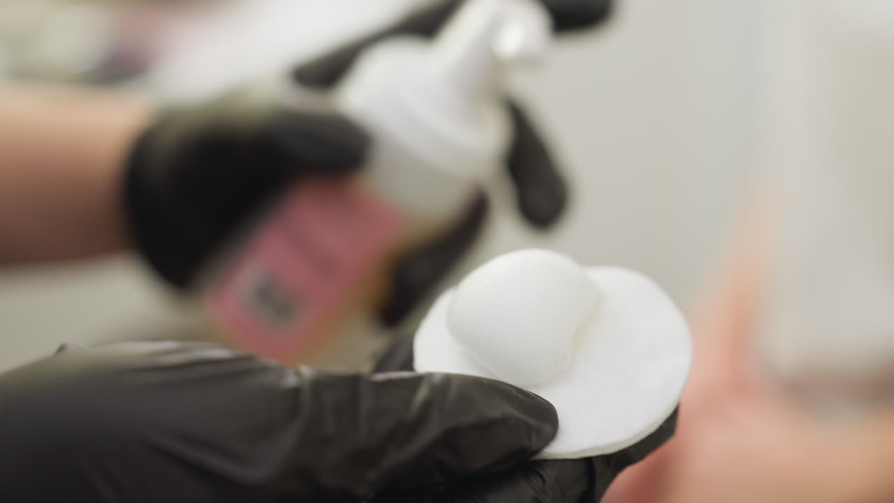 Beautician wearing black gloves dispenses foam cleanser onto cotton pad while preparing to clean eyebrow using brush, demonstrating skincare precision and hygiene in bright salon environment
