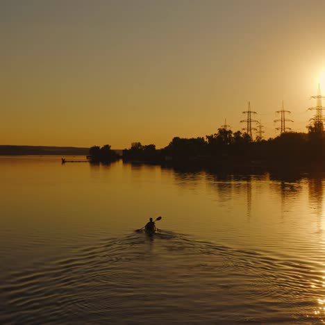 silhouette of a factory near the river. man in a boat roaming with oars at sunset. high voltage electric towers near the water against the setting sun.