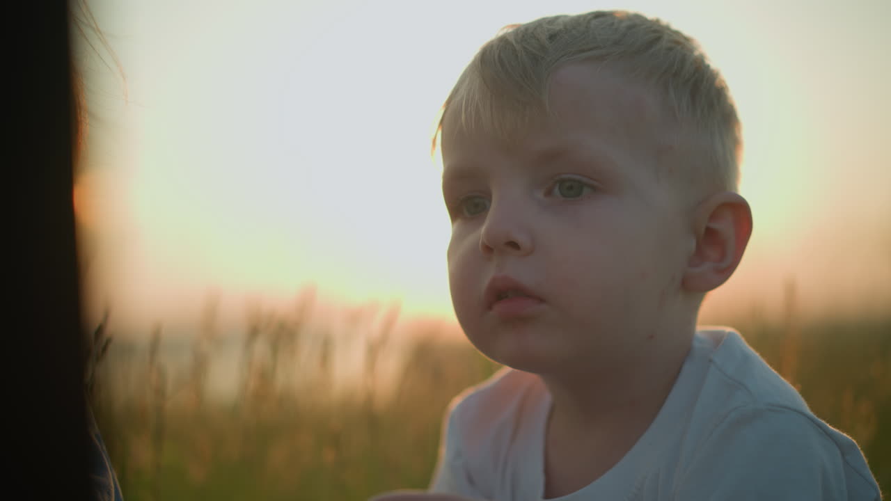 Close-up of a young boy in a white shirt, looking contemplative and slightly sad, against the serene backdrop of a grassy field at sunset