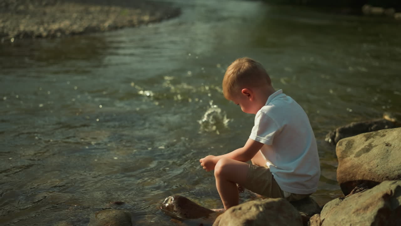 Young boy plays in park throwing stones into shallow river water at sunset. Little child relishes atmospheric evening with amazing landscape and clear water