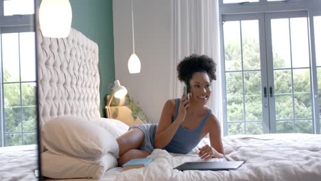 mujer biracial feliz acostada en la cama y hablando en el teléfono inteligente en el dormitorio, cámara lenta