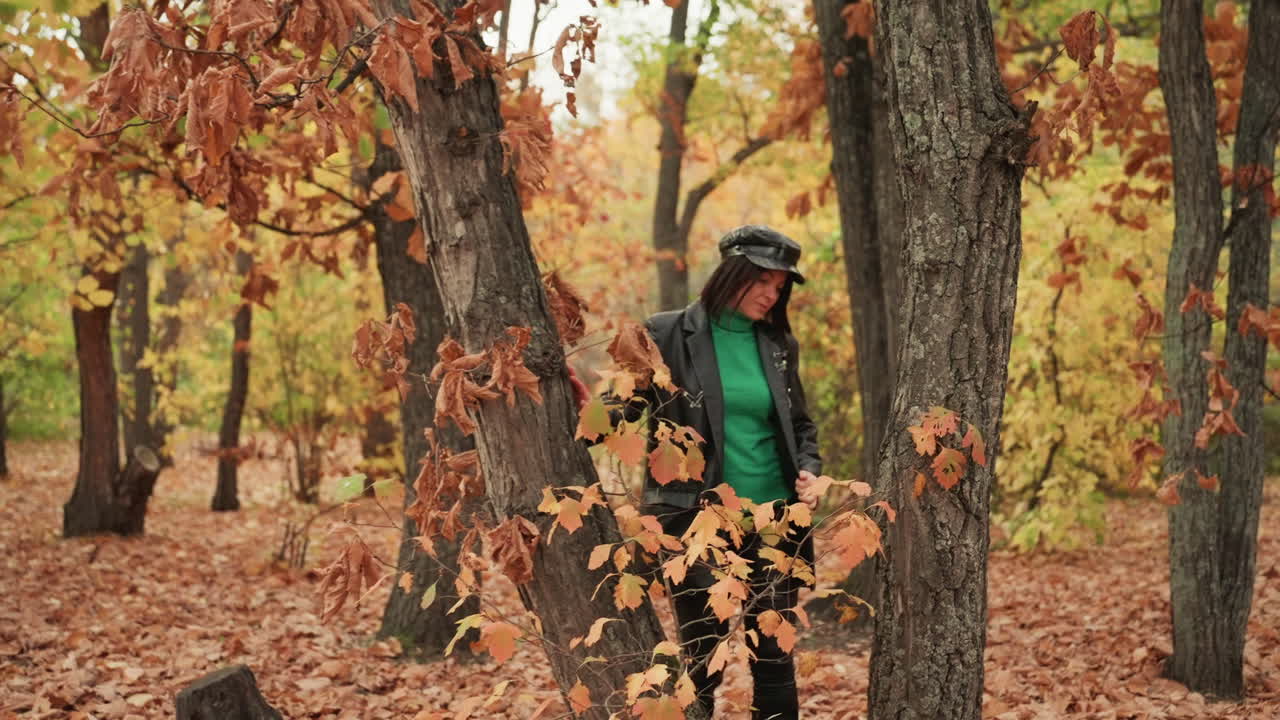 Žena opřená o dub v kožené bundě a kapuci, zamyšlená rockerka s klidným pohledem a rustikálním pozadím listoví, drsná pouliční atmosféra setkávající se s přírodou, samotou a reflexí