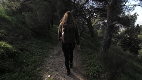 young woman walks on path among trees and grass on sunny day, looks back at camera and smiles, gimbal following