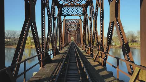 Drone-flight-on-a-rusty-train-bridge-over-a-river-with-blue-sky-on-a-sunny-day,-dolly-shot