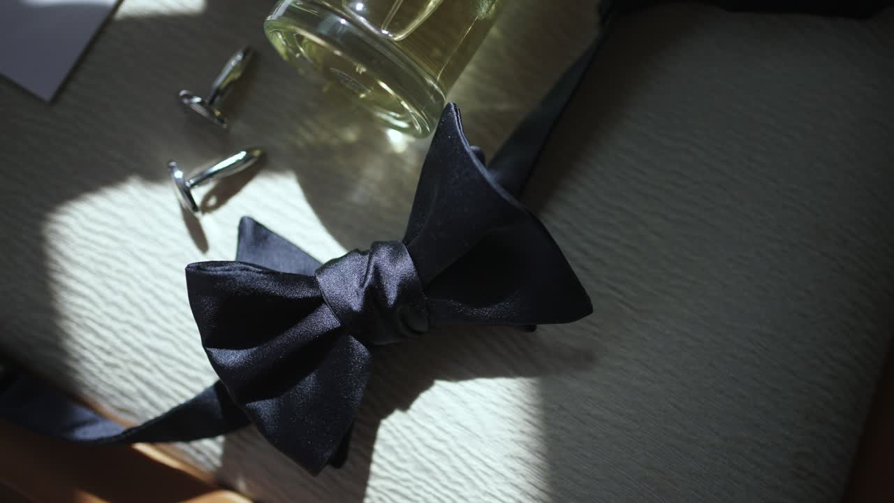black bow tie next to cufflinks and cologne bottle on textured surface lit by soft sunlight