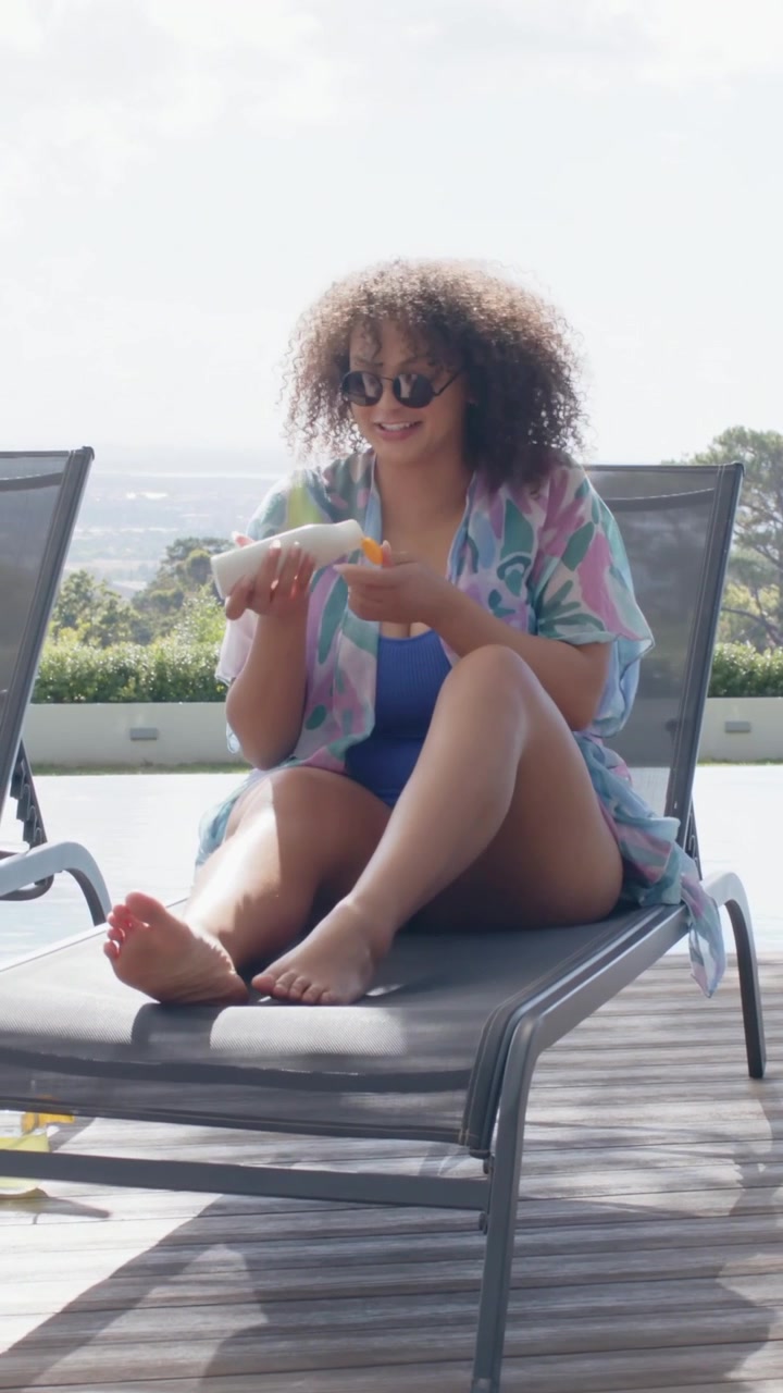Video verticale: Guardando a sinistra, donna in costume da bagno blu che prende la crema solare in piscina, spalmando le gambe per il sole.